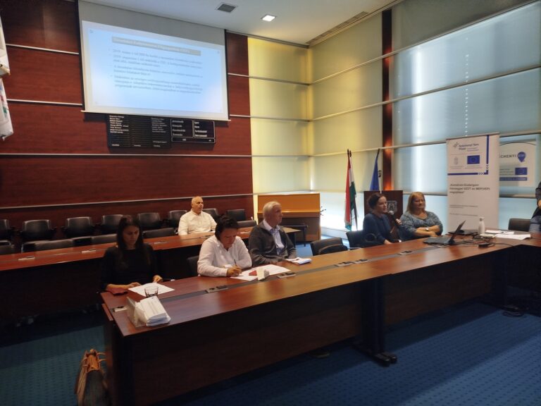 Munkacsoporti egyeztetés zajlik a vármegyeháza tanácstermében, résztvevők jegyzetelnek és számítógépet használnak, háttérben kivetített képernyő.