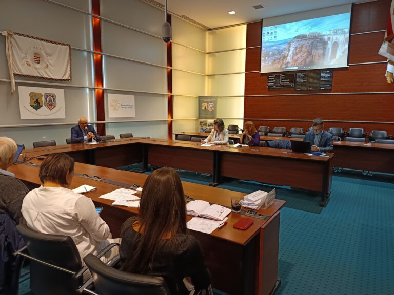 Munkacsoporti egyeztetés zajlik a vármegyeháza tanácstermében, résztvevők jegyzetelnek és számítógépet használnak, háttérben kivetített képernyő.