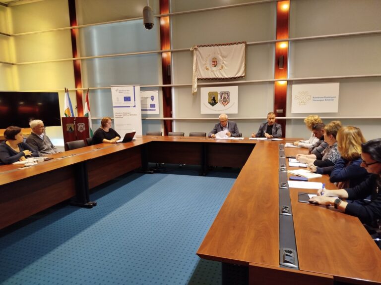 Felzárkózási Fórum ülés Komárom-Esztergom vármegyei tanácsteremben, körasztalos elrendezésben, háttérben hivatalos jelképek.